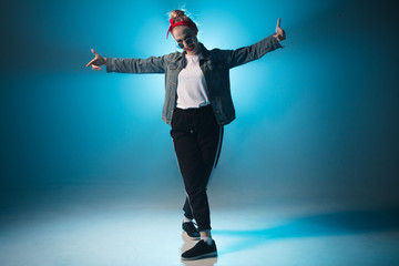 Young woman dressed in street fashion wear dancing hip-hop style over studio blue light background with flare effects