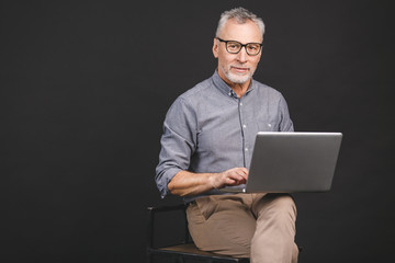 Grandfather using modern technologies. Smiling old businessman in eyeglasses using laptop computer isolated over black background.