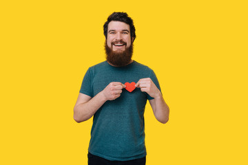 Handsome happy bearded man looking at the camera, holding a red heart upon his chest on yellow background.