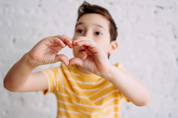 Happy tweens boy in yellow t-shirt making heart by hands on the white wall background, selective focus