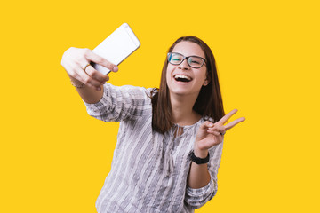 Charming young woman take selfie on front camera smartphone showing v-sign isolated on yellow wall background