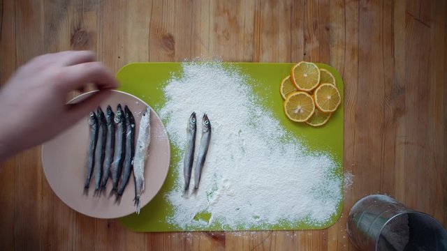 Top view of a man's hands covering a capelin with flour on a wooden table, the process of making a small silver fish, preparation for frying in oil in a pan.