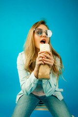 young beautiful blonde girl eating Fast food, a woman with an appetite eating Shawarma on a blue background, in denim clothes and glasses