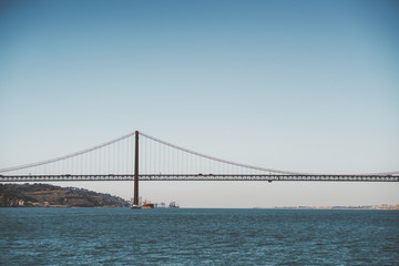 A view from the water of a huge suspension bridge "Ponte 25 de Abril" over the river Tagus in Lisbon, Portugal on a warm morning day with the horizon in the background
