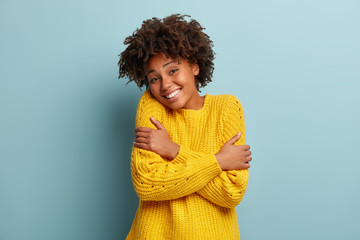 Headshot of pleased dark skinned woman hugs herself, smiles from pleasure, likes new yellow sweater she bought, has Afro hairstyle, crosses hands, recalls romantic moment, isolated on blue wall - obrazy, fototapety, plakaty