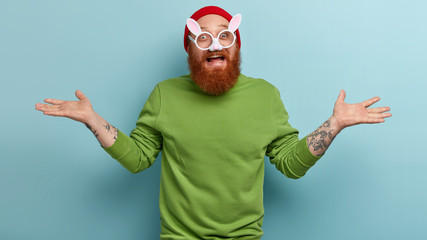 Horizontal shot of cheerful bearded unshaven man shruggs shoulders, has funny look, wears rabbit...
