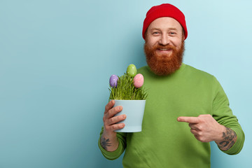 Glad foxy hipster male happy to bring colored Easter eggs, points at pot where they grow, being in festive mood, dressed in bright fashionable clothes, stands over blue wall, empty space away - Powered by Adobe