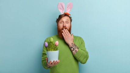 Bearded Caucasian man has astonished expression, covers mouth with hand, holds pot with colored Easter, acts as bunny who brings presents to chidren, stands over blue background. Rabbit ears on head