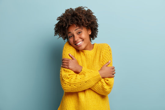 Headshot of pleased dark skinned woman hugs herself, smiles from pleasure, likes new yellow sweater she bought, has Afro hairstyle, crosses hands, recalls romantic moment, isolated on blue wall