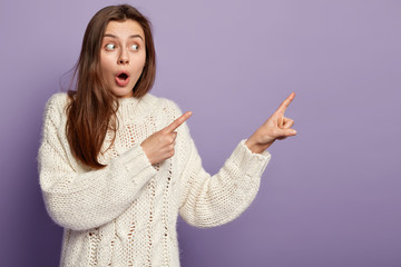 Portrait of expressive emotional woman points away with fore fingers, looks aside with opened...