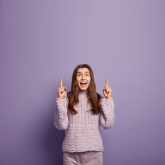 Positive European young woman points both fore fingers above, shows cool place upstairs, laughs, being amused and pleased, wears oversized winter warm sweater, isolated over purple background.