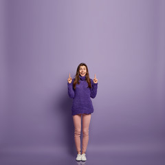 Cheerful lovely woman with satisfied expression, notices something wonderful upwards, points fore fingers above, wears knitted sweater, poses over purple studio wall with empty space for promotion
