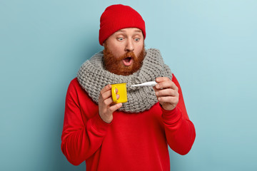 Photo of ill bearded man stares at thermometer, surprised to have high temperature, has flu, wears red headgear, scraf, drinks hot tea, isolated over blue background. People and disease concept
