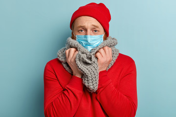 Photo of sick man has infection, flu, wears medical mask, suffers from contagious disease, treats cold, has knitted scarf around neck, wears red headgear and jumper, isolated on blue background