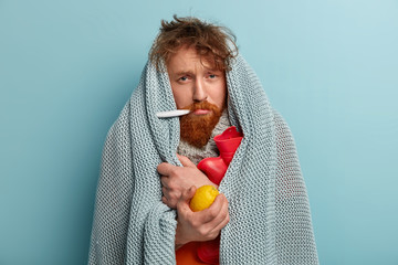 Redhead young man with bristle fights against flu and fever, holds lemon to enrich vitamins, has...