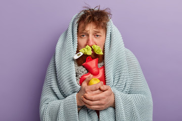 Medical treatment concept. Bearded ill foxy man wrapped in coverlet, holds hot water bottle, carries lemon, has thermometer in mouth, trembles from fever and freezing, stands over purple wall