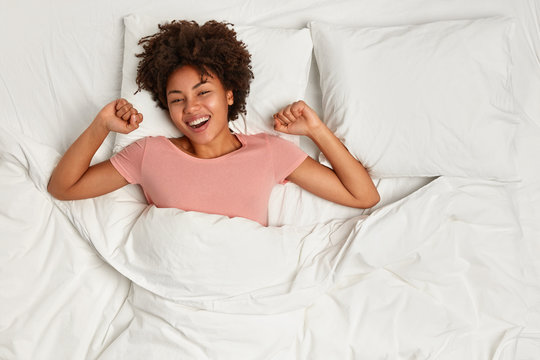 Photo of cheerful satisfied lovely young woman with curly hair wakes up in morning optimistic, stretches in bed with happy expression, has day of relaxation during weekend. Time for rest concept