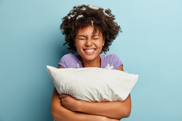 Photo of joyful impressed cheerful woman smiles broadly, has pillow fight, feathers stuck in curly...