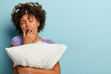 Sleepy tired young Afro American woman covers mouth with hand, yawns after sleep, embraces white pillow, has feathers in head, poses over blue background with blank space for your promotion.