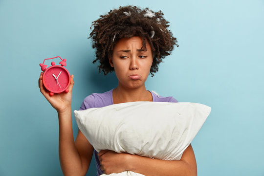 Photo of sad Afro American young woman purses lower lip, carries pillow, holds alarm clock, unhappy to have not enough sleep, isolated over blue background, upset wake up early, needs more time