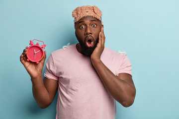 Frightened young black man opens mouth widely, keeps hand on cheek, carries alarm clock, wears eyemask and casual t shirt, isolated over blue background, being late, impressed early in morning - obrazy, fototapety, plakaty