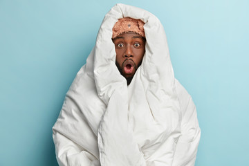 Photo of stupefied dark skinned man wrapped in white blanket, feels cold in room, cant get asleep,...