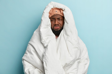 Gloomy Afro American guy wrapped in soft duvet, wears eyemask, has unhappy look, cries in dissatisfaction, doesnt want to get up early at weekend, isolated on blue background. Sullen sleepy man