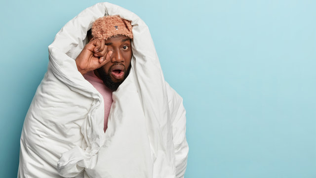 Its difficult to wake up early. Dark skinned sad man rubbs eyes, wants to sleep, wrapped in white blanket, has sleepy tired look, stands over blue wall with free space aside. Awakening concept