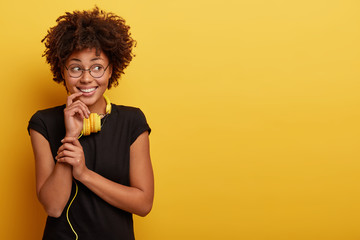 Pleased impressed dark skinned woman looks aside, has dreamy expression, pleasant smile, listens music with modern headphones, dreams about something, isolated on yellow wall with copy space