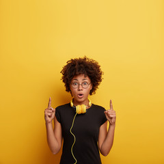 Indoor shot of shocked young Afro American woman listens to stream of music, uses music app, points...