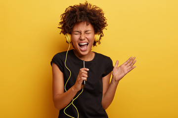 Amused black young woman with curly hair, shakes hands, enjoys cool music, holds smart phone,...