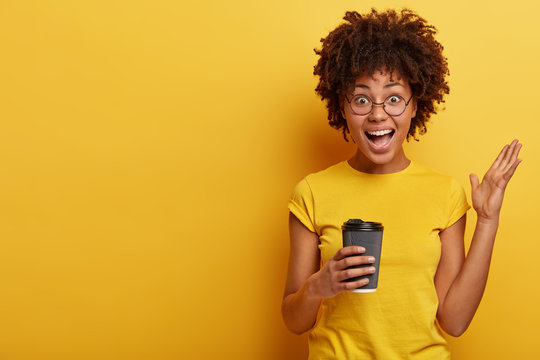 Happy optimistic black woman gestures actively, drinks hot beverage, talks emotionally with friends, wears bright t shirt in one tone with background has nice cup of coffee boosts energy with caffeine