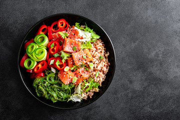 poke bowl of brown rice, trout or salmon fillet, Chuka Seaweed Salad, sesame and fresh vegetables on dark background, top view, copy space, healthy food trend