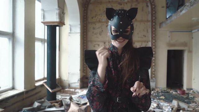 Young beautiful girl stands on drum among the garbage in a ruined building. She wears a leather sword, and a cat's mask made of leather. She dances, depicting a cat, smiling, close-up
