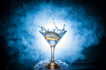 Martini cocktail glass in hand splashing on dark toned smoky background or colorful cocktail in glass with splashes and olives. Party club entertainment. Mixed light. Selective focus