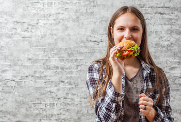 portrait of young teenager brunette girl with long hair eating burger. Girl trying to eat fast food on gray wall background