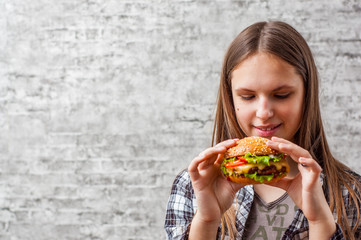 portrait of young teenager brunette girl with long hair eating burger. Girl trying to eat fast food on gray wall background