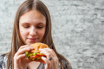 portrait of young teenager brunette girl with long hair eating burger. Girl trying to eat fast food on gray wall background