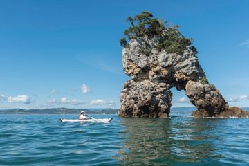 Adult, female kayaker paddling past an offshore natural arch in a rock stack with trees growing on top. On a sunny, summer's day in Coromandel, New Zealand.