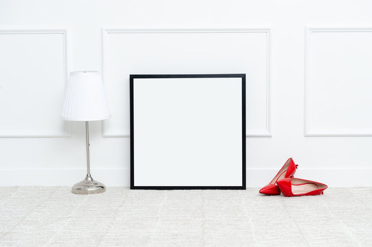 There are beautiful high heels and various objects near the rectangle blank picture frame in front of white wall on the carpet living room.