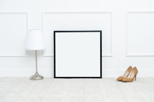 There are beautiful high heels and various objects near the rectangle blank picture frame in front of white wall on the carpet living room.