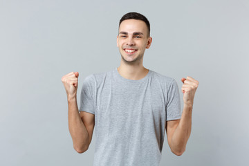 Fototapeta premium Portrait of cheerful young man in casual clothes looking camera, clenching fists like winner isolated on grey wall background in studio. People sincere emotions, lifestyle concept. Mock up copy space.