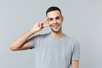 Fototapeta premium Portrait of handsome smiling young man in casual clothes looking camera showing victory sign isolated on grey wall background in studio. People sincere emotions, lifestyle concept. Mock up copy space.