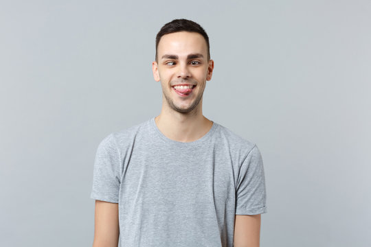 Portrait of crazy loony funny young man in casual clothes with beveled eyes showing tongue isolated on grey wall background in studio. People sincere emotions, lifestyle concept. Mock up copy space.