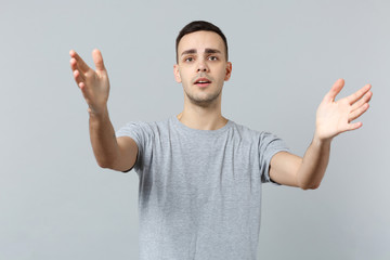 Portrait of displeased young man in casual clothes stand with spreading outstretching hands isolated on grey wall background in studio. People sincere emotions, lifestyle concept. Mock up copy space.