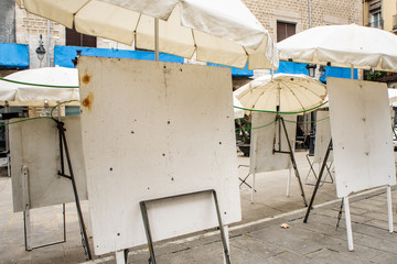 Empty dirty metal white easels on the street under white umbrellas. Street artists are gone for weekend. Selll paintings in the streets.