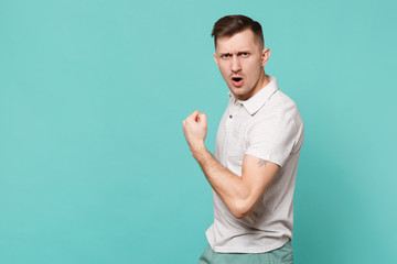 Portrait of strong young man in casual clothes standing, showing biceps, muscles isolated on blue turquoise wall background in studio. People sincere emotions, lifestyle concept. Mock up copy space.