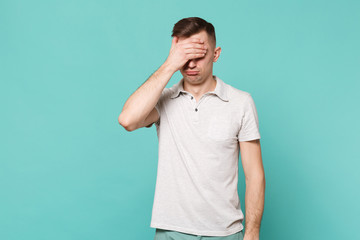 Portrait of offended young man in casual clothes covering eyes with palm isolated on blue turquoise wall background. People sincere emotions, lifestyle concept. Mock up copy space. Advertising area.