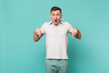 Surprised young man in casual clothes keeping mouth open, pointing index fingers down isolated on blue turquoise background in studio. People sincere emotions, lifestyle concept. Mock up copy space.