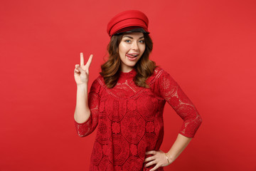 Obraz na płótnie Canvas Portrait of pretty cute young woman in lace dress, cap showing tongue and victory sign isolated on bright red wall background in studio. People sincere emotions lifestyle concept. Mock up copy space.